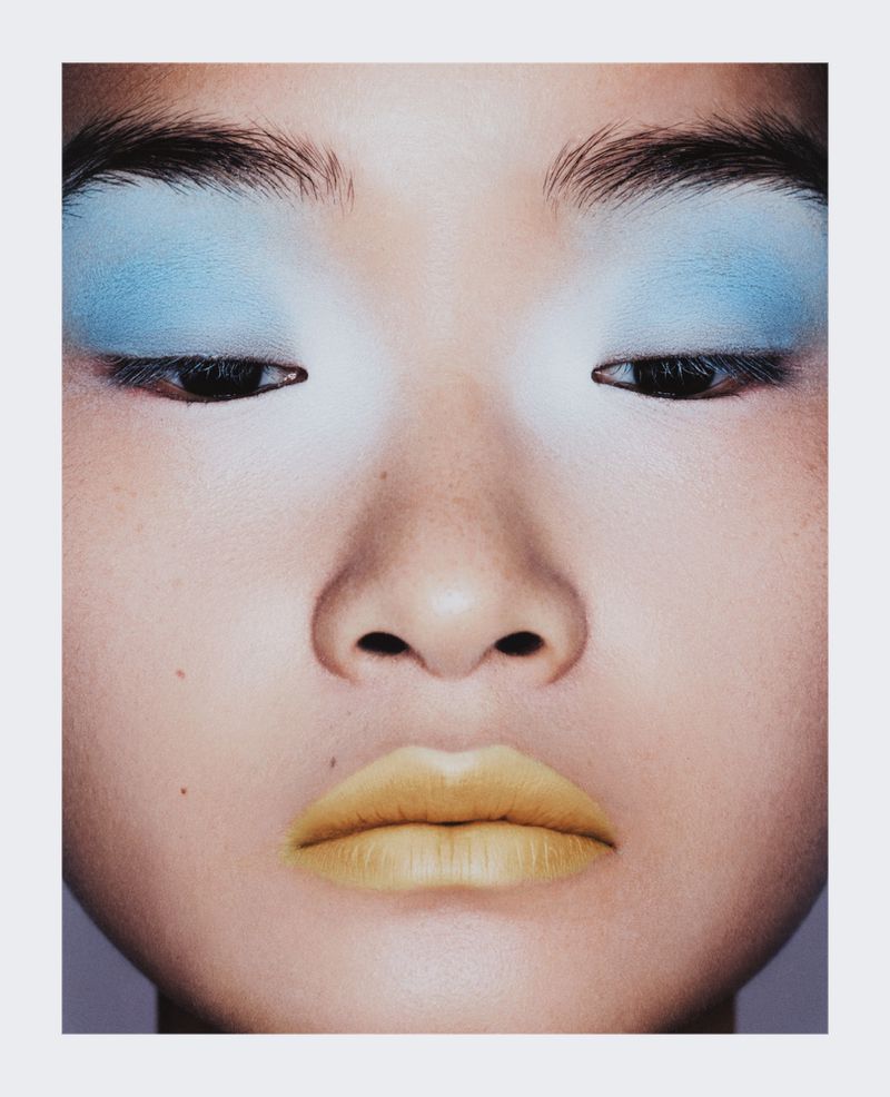 Close-up portrait of a woman with bright blue eye shadow and light yellow lipstick against a plain off-white background.