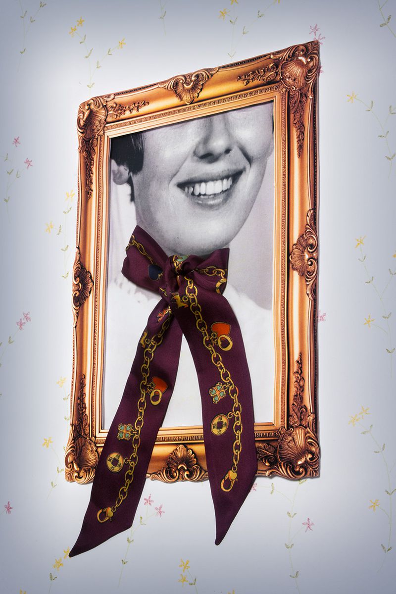 Framed portrait of a smiling woman with a patterned maroon scarf tied around the frame and neck, against a floral wallpaper backdrop.