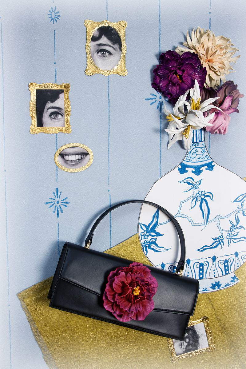 Stylish black leather clutch handbag with a burgundy flower accent, set against a light blue patterned backdrop with retro eye artwork and a floral vase.