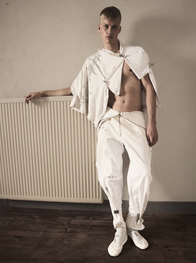 Young adult man in white avant-garde fashion poses in front of a radiator in an indoor setting, showcasing modern style.