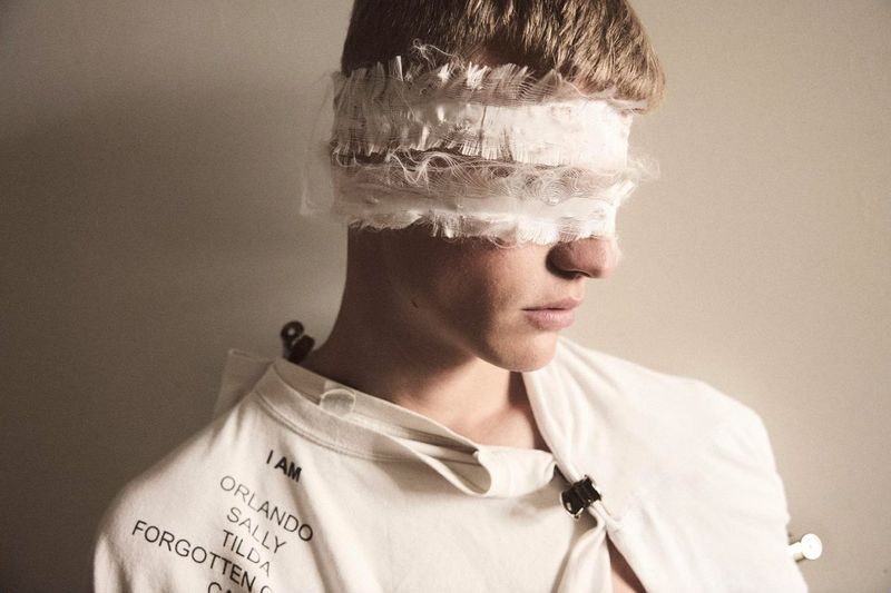 Young adult male portrait with a layered white fabric blindfold; close-up shot with neutral background, indoor setting, wearing a white garment.