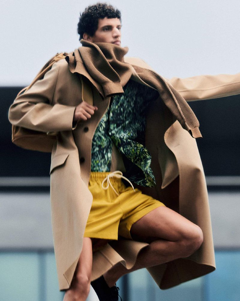 Man in motion wearing a tan trench coat, brown sweater, and yellow shorts, captured in an editorial fashion style against a neutral backdrop.
