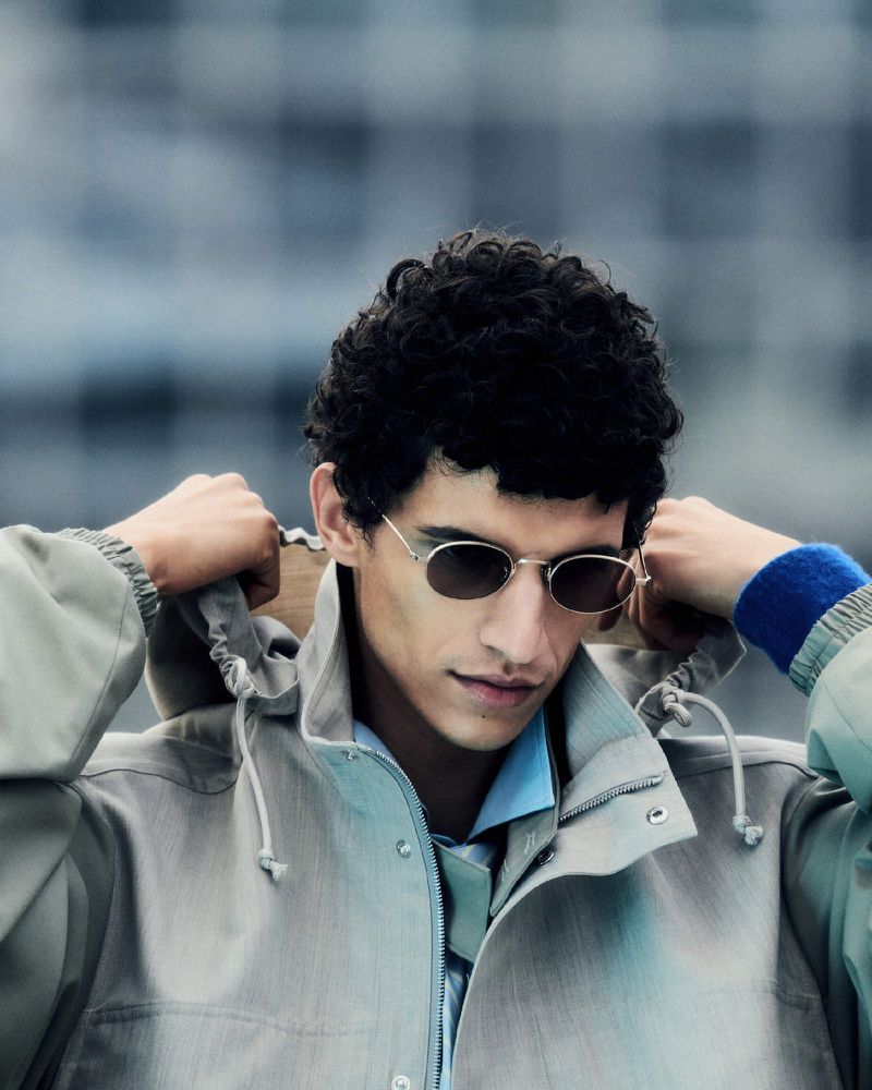 Portrait of a young adult male adjusting the hood of a light tan jacket while wearing sunglasses, captured in a medium shot.