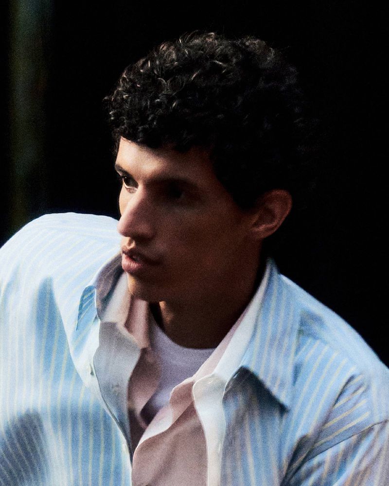 Close-up portrait of a man with curly dark hair wearing a light blue and white striped shirt layered over a white t-shirt.
