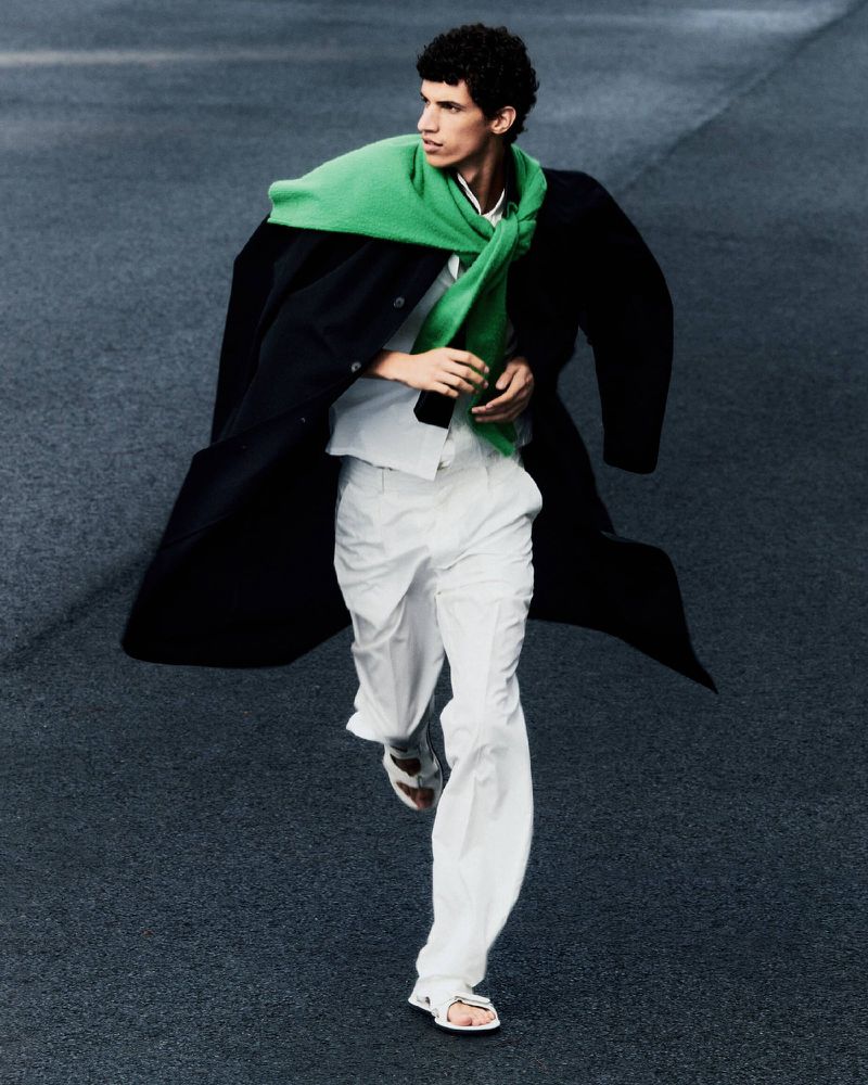 A young adult man in a black coat, white pants, and sandals is captured in motion with a green scarf against a dark asphalt background.