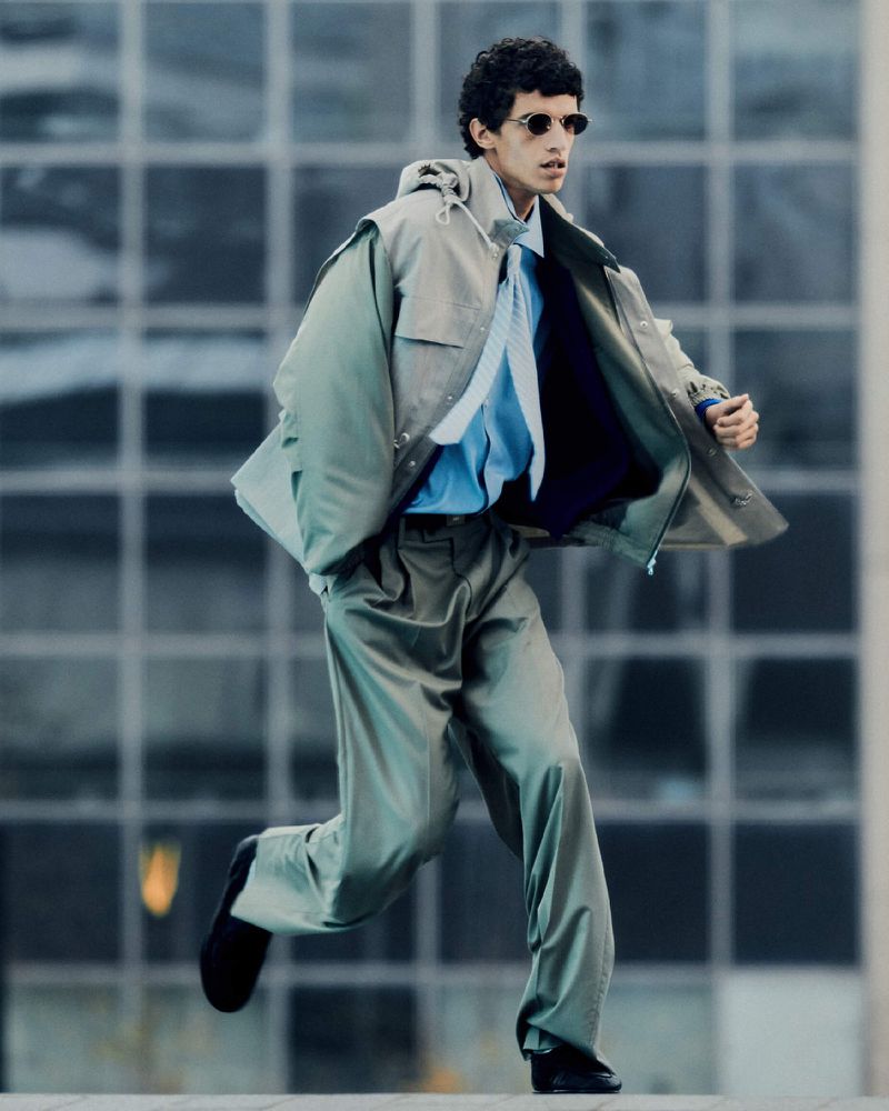 A man running outdoors wearing a light grey oversized suit, pale blue shirt, and sunglasses against a blurred building backdrop.