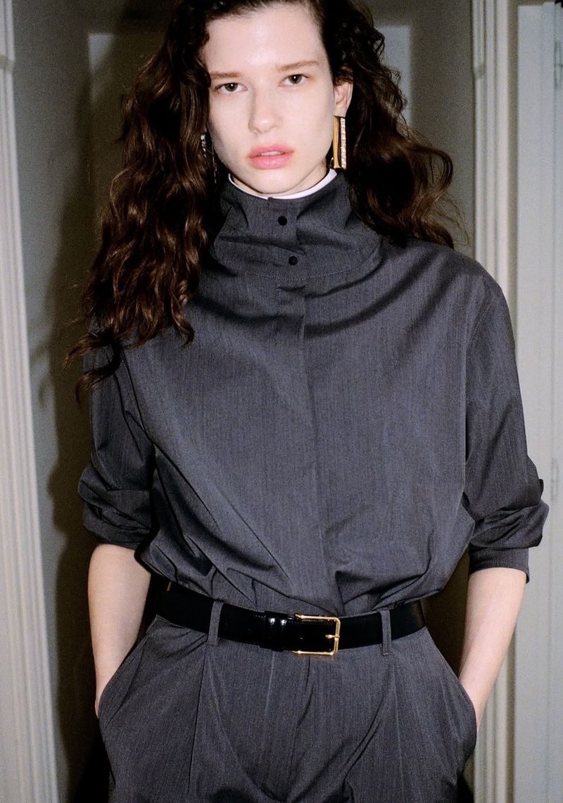 Fashionable woman in a gray pinstripe suit stands indoors, accessorized with a black belt and gold earrings, showcasing contemporary style.