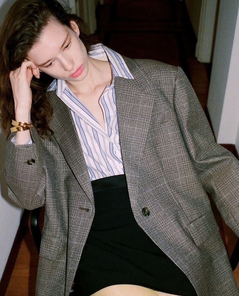 Adult female in an oversized blazer and mini skirt poses indoors, wearing gold bangles and a striped shirt in a medium shot.