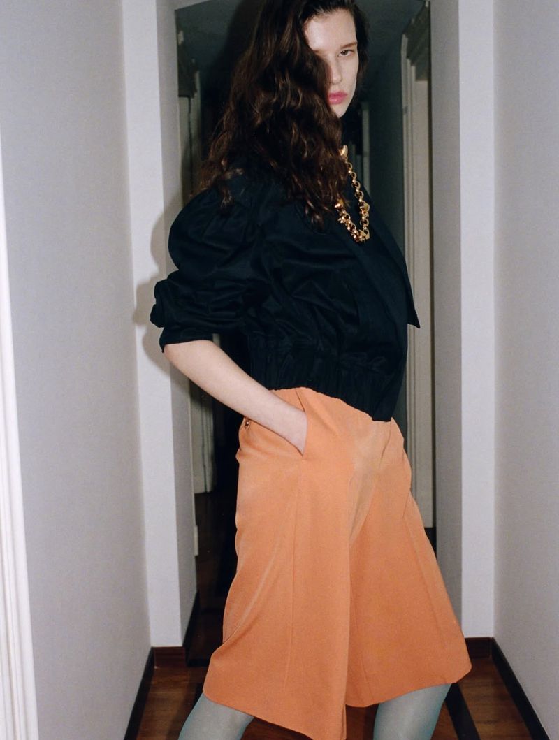 A young woman poses indoors wearing a black jacket, terracotta-colored culottes, and a gold necklace, standing in a hallway.