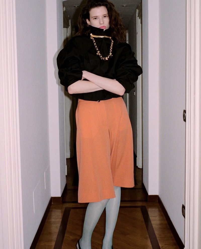 A person standing indoors with arms crossed, wearing a black top, orange shorts, sheer tights, and flats with a gold necklace.