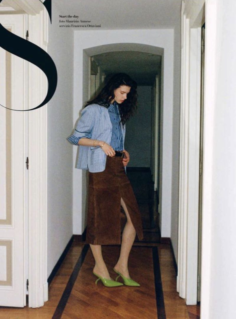 A stylish woman in a striped shirt, blazer, and brown skirt stands indoors. The woman is posing near a doorway with a hallway.