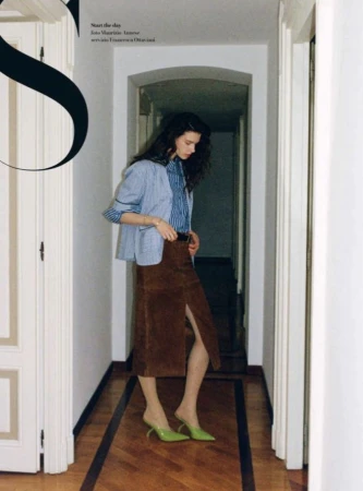 A stylish woman in a striped shirt, blazer, and brown skirt stands indoors. The woman is posing near a doorway with a hallway.