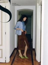 Sandra Murray wearing a blue shirt jacket, brown suede slit skirt, and lime green pointed mules in an interior hallway.
