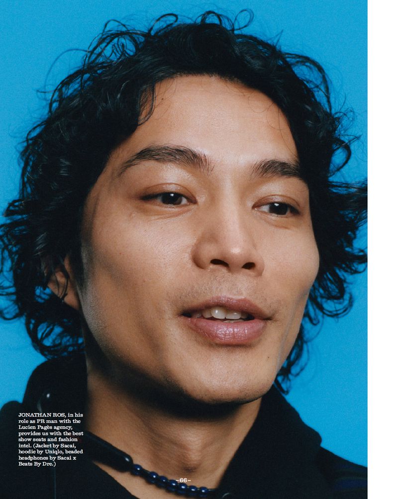 Close-up studio shot of a curly-haired adult male wearing a black top and beaded necklace against a bright cyan background.
