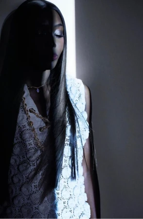 Woman in a sleeveless, white lace top stands in a shadowed space with a beam of bright light accenting her long brown hair.