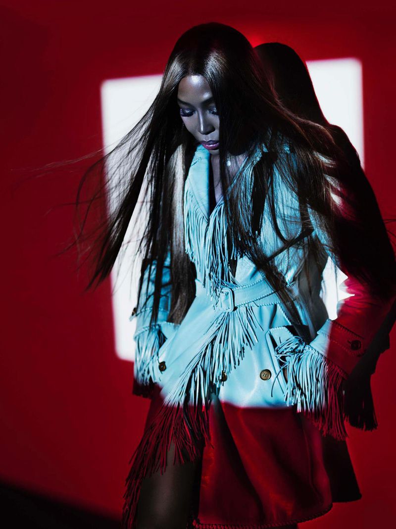 A fashion shot of a woman with long dark hair wearing a fringed light blue jacket and red skirt against a vivid red backdrop.