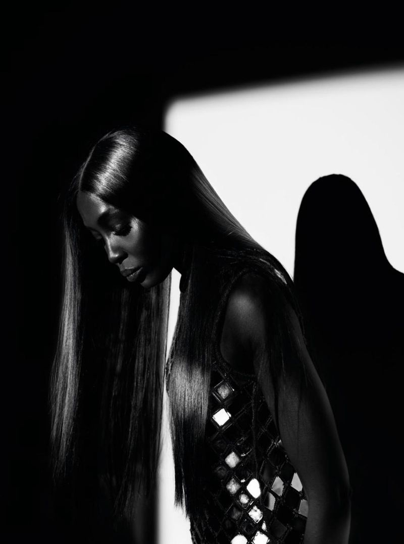 Black and white portrait of a Black female model with long straight hair in a sleeveless dress, posing in front of a light panel.