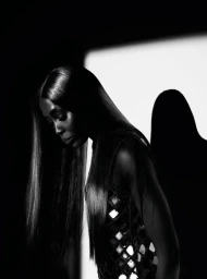 Black and white portrait of supermodel Naomi Campbell looking down, wearing a mirror-embellished top against dramatic studio lighting.
