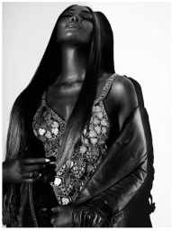 Naomi Campbell posing in an embellished patterned top and draped leather jacket against a light studio backdrop.
