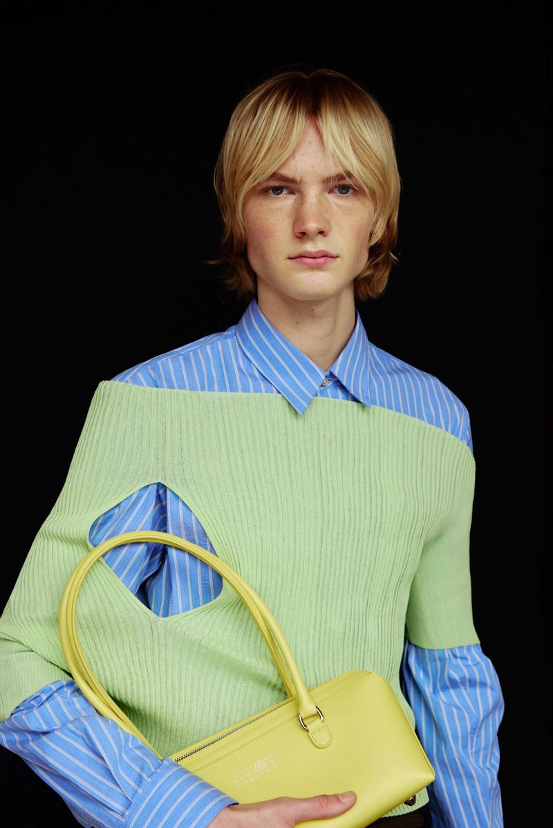 Young man in a blue and white striped shirt layered with a green sweater, holding a yellow bag against a stark black backdrop.