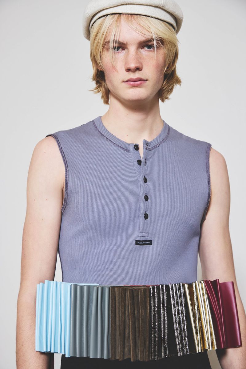 Fair-skinned male model wearing a cap and sleeveless shirt, holding color swatches, studio shot against a white backdrop. Fashion-forward portrait.
