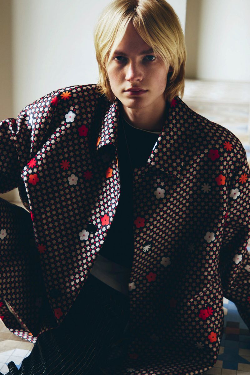 Blonde haired model poses in a patterned jacket against a neutral backdrop, showcasing a relaxed, stylish aesthetic.