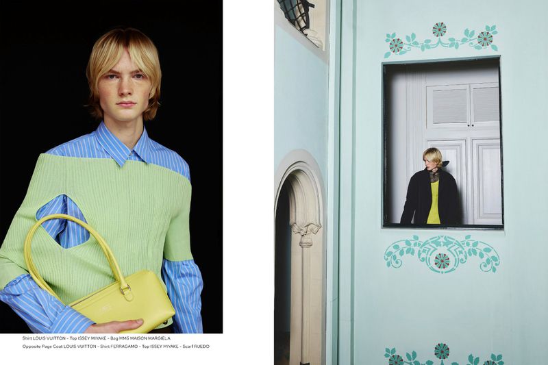 Fashion editorial shot of a slender young man wearing a blue striped shirt and cut-out green sweater, holding a yellow bag, in a photo studio.