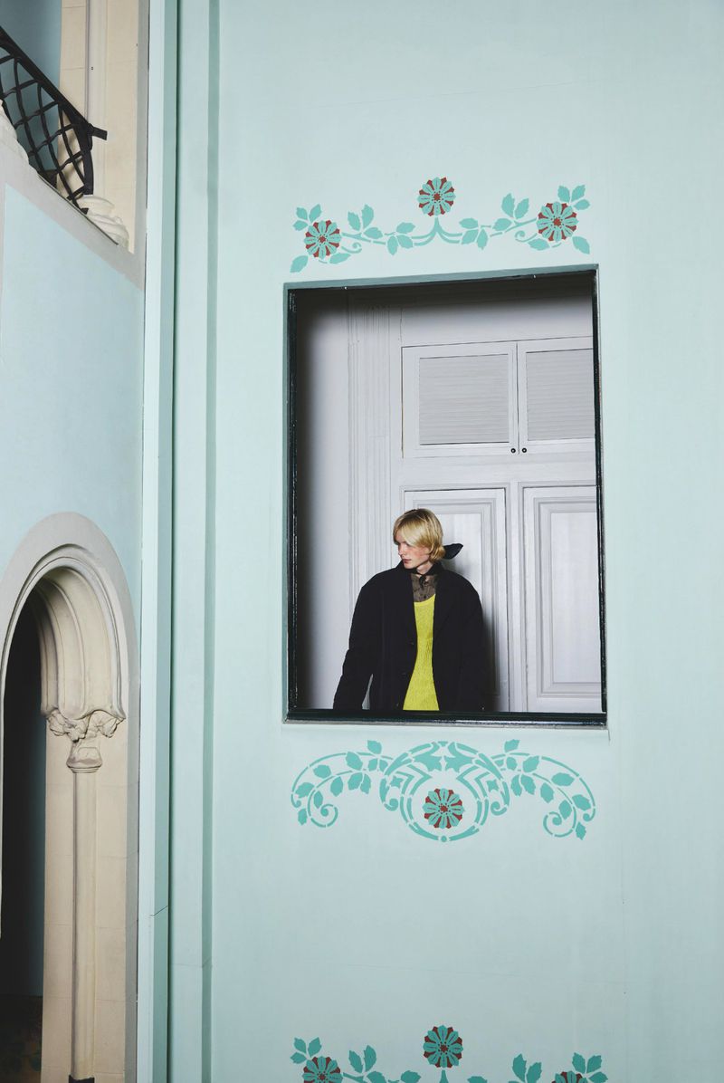 Woman with short blonde hair in black jacket and yellow shirt stands in window of light blue room with floral decorations.
