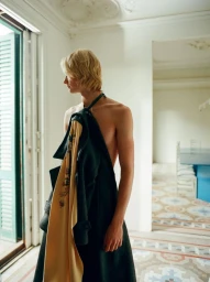 Model Leander Straub posing shirtless draped in a dark green coat with yellow lining inside a bright sunlit room with tiled floors.