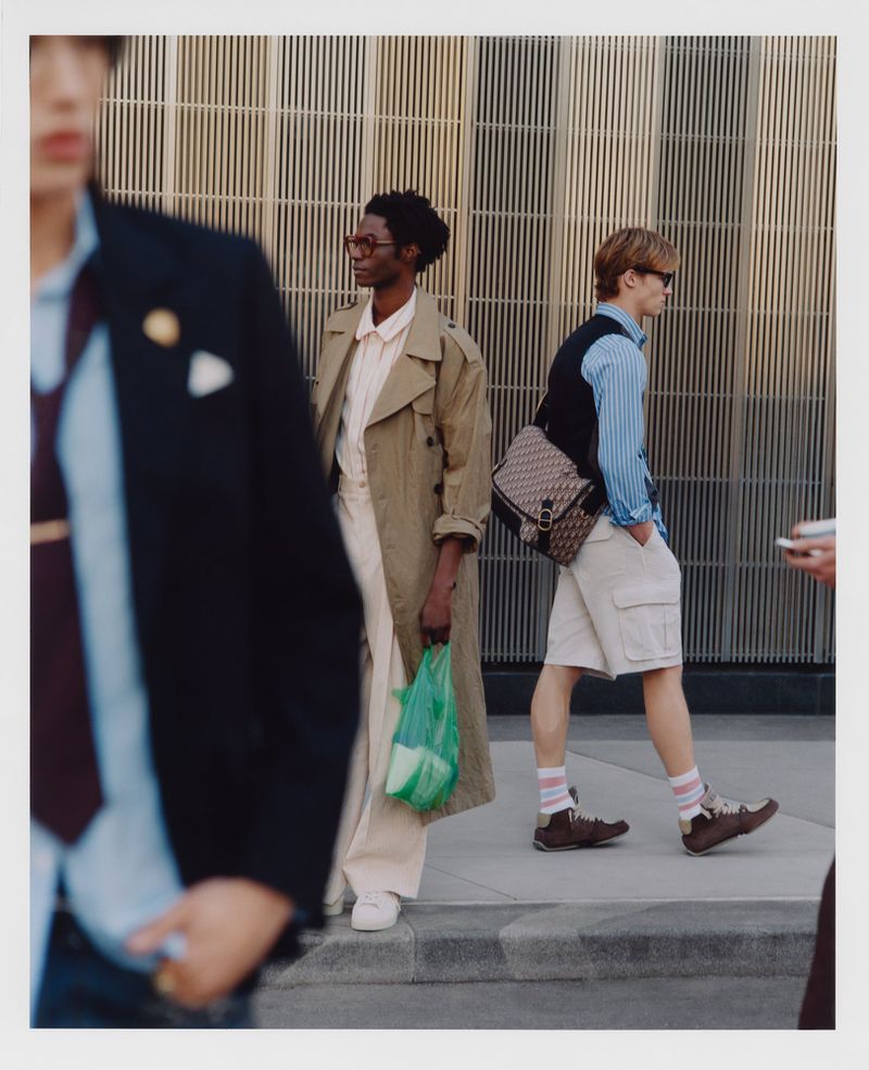 Three stylish individuals walking on a city sidewalk against a modern building. One carries a green plastic bag and another a designer bag.