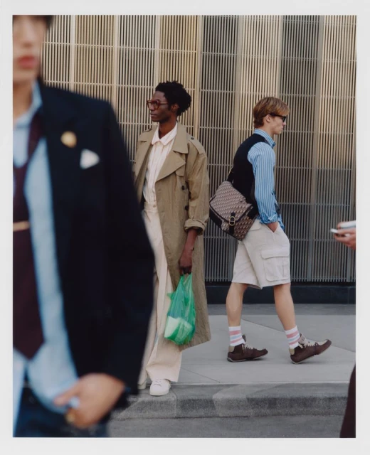 Three stylish individuals walking on a city sidewalk against a modern building. One carries a green plastic bag and another a designer bag.