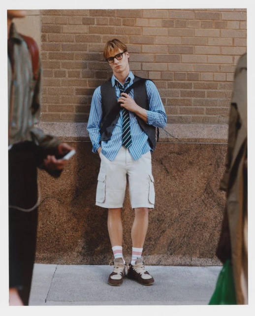 A young man wearing a striped shirt, vest, tie, and shorts stands in an urban scene. People are walking past him in the background.