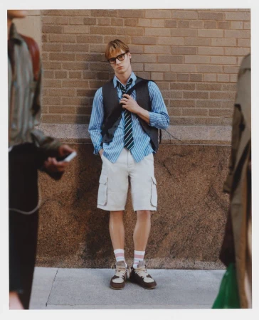 A young man wearing a striped shirt, vest, tie, and shorts stands in an urban scene. People are walking past him in the background.