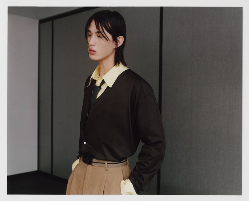 Portrait of a young adult with long dark hair wearing a brown shirt and khaki pants, posed against a gray wall with natural lighting.