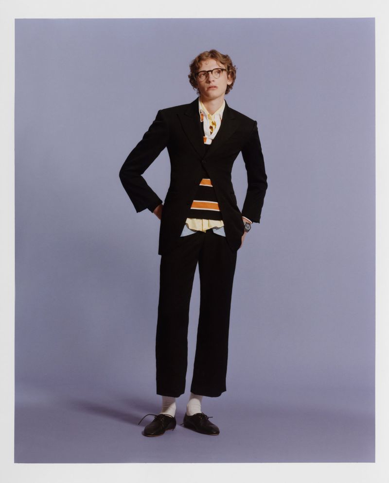 A man in a black suit, patterned shirt, and eyeglasses poses against a lavender backdrop in a fashion studio portrait with eye-level angle.