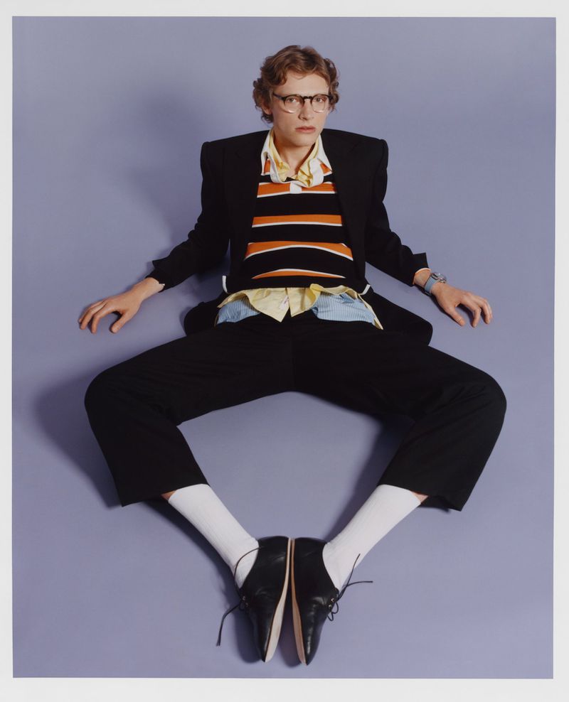 Full studio shot of a person with short hair wearing glasses, a striped shirt, black blazer and pants, white socks, and black dress shoes.