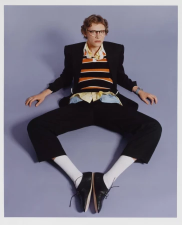 Full studio shot of a person with short hair wearing glasses, a striped shirt, black blazer and pants, white socks, and black dress shoes.
