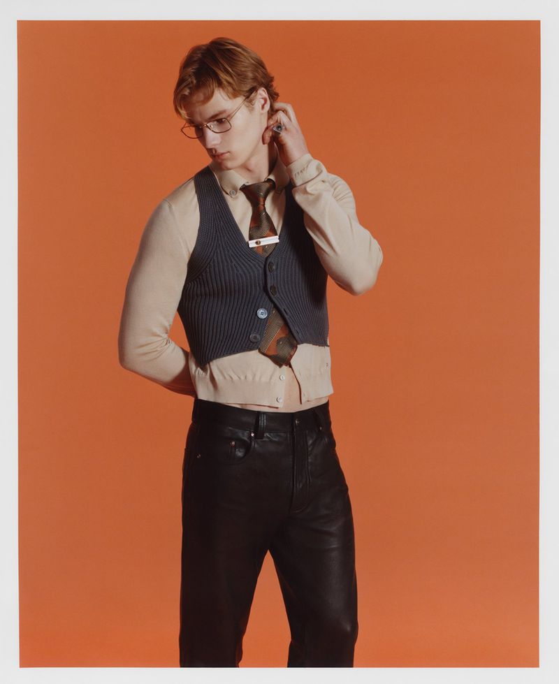 Adult male wearing glasses, a vest, a collared shirt, and dark pants poses in front of a solid orange backdrop, looking stylish and contemplative.