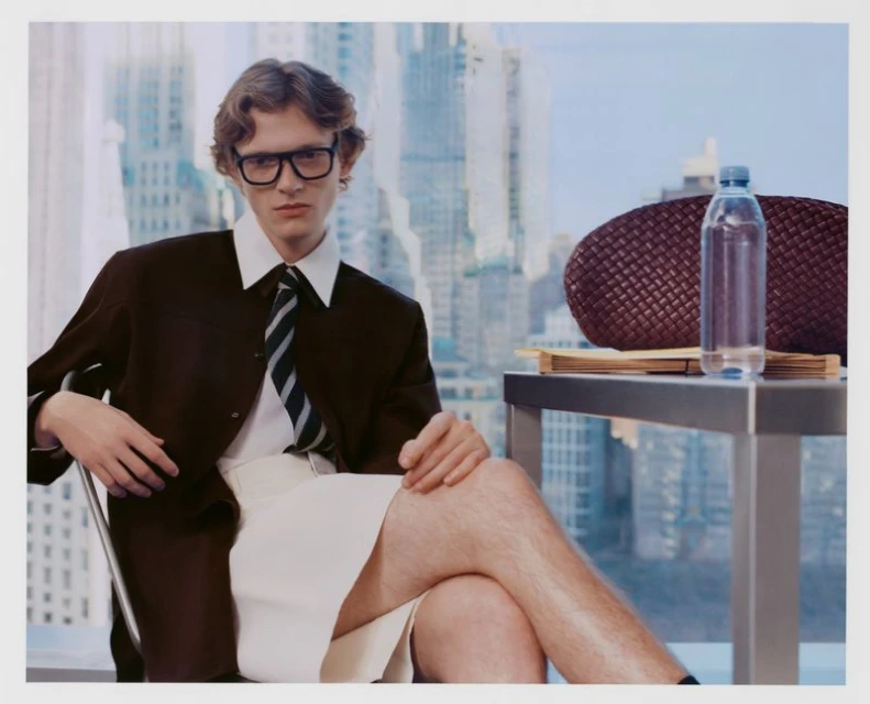 Man in brown suit poses indoors by window overlooking a city, sitting at a desk with water bottle and leather bag.