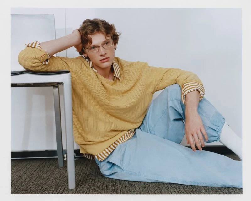 Young adult with auburn hair sits on floor in light-blue pants, yellow top, eyeglasses, leaning on metal-framed chair, indoor studio shot.