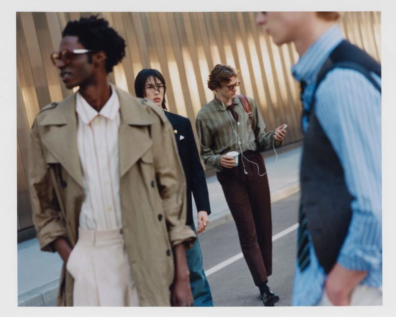 Four young adults stand on a sidewalk in fashionable outfits near a textured facade, with one holding a coffee cup and another an iPhone.