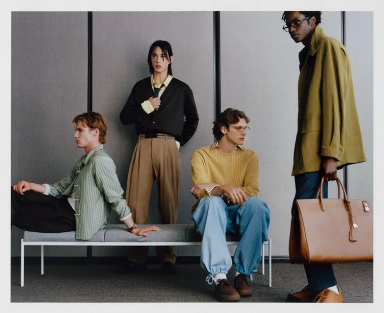 Four stylish men in modern, relaxed fashions pose in a neutral indoor setting, showcasing contemporary clothing and accessory trends.