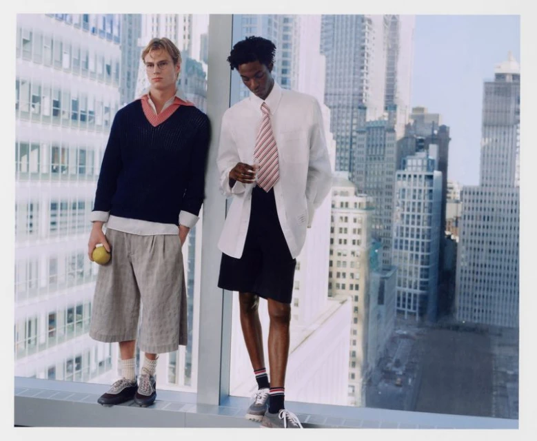 Two men pose against a window overlooking a city. One man wears a navy sweater, the other a white shirt and jacket with shorts.