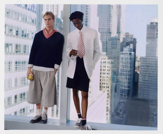 Two men pose against a window overlooking a city. One man wears a navy sweater, the other a white shirt and jacket with shorts.
