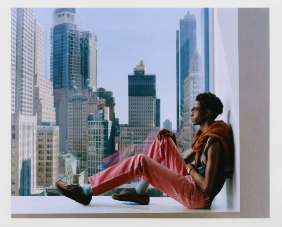 Young Black man in glasses and pink pants sits at a window in a modern high-rise apartment with a cityscape view.