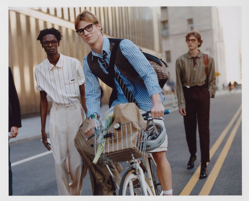 Three stylish men pose on a city street, one riding a bicycle with groceries, showcasing fashion, city life, and urban commuting.