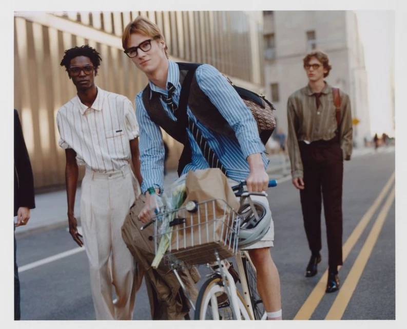 Three stylish men pose on a city street, one riding a bicycle with groceries, showcasing fashion, city life, and urban commuting.