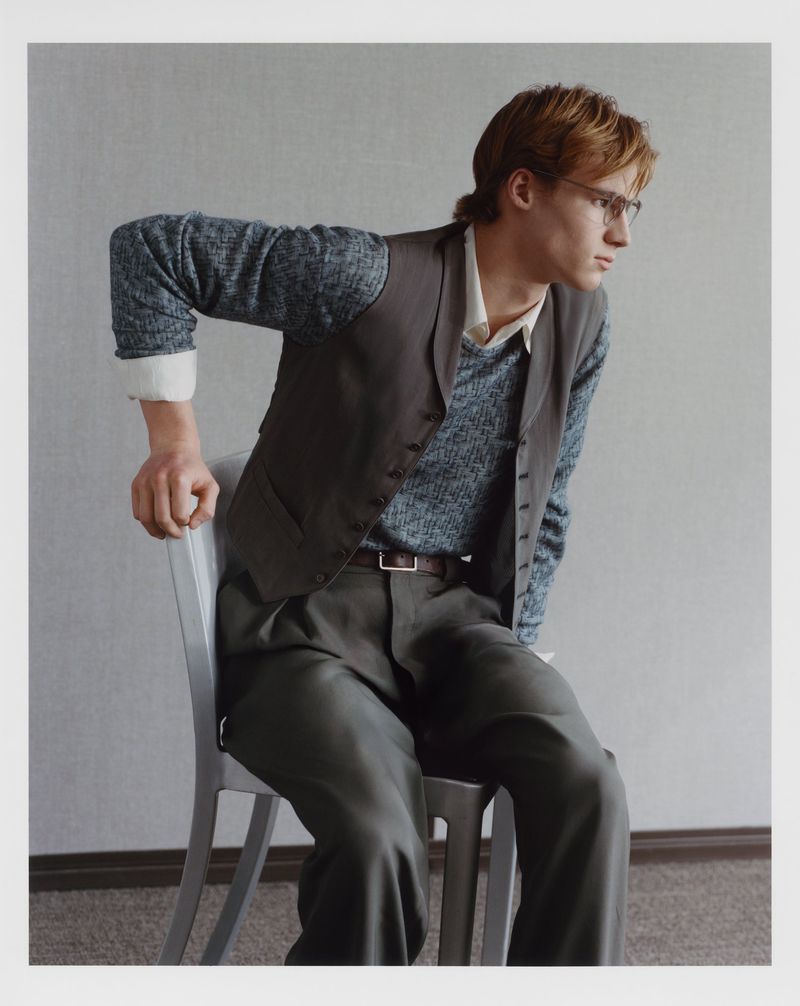 Portrait of a young adult man with blond hair and glasses, wearing a vest, shirt, and pants, sitting on a chair in a photo studio.