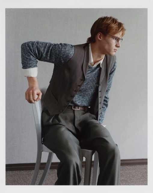 Portrait of a young adult man with blond hair and glasses, wearing a vest, shirt, and pants, sitting on a chair in a photo studio.