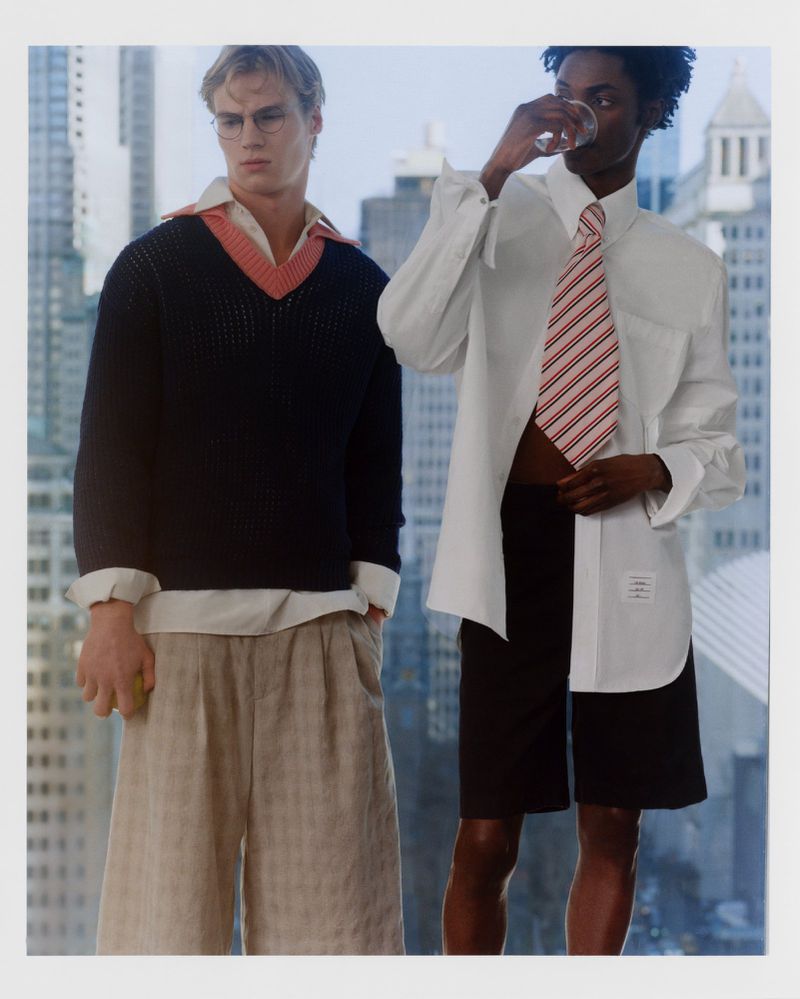 Two fashion models pose outdoors in front of a city backdrop. One is drinking water; both wear shirts, shorts and ties in a retro style.
